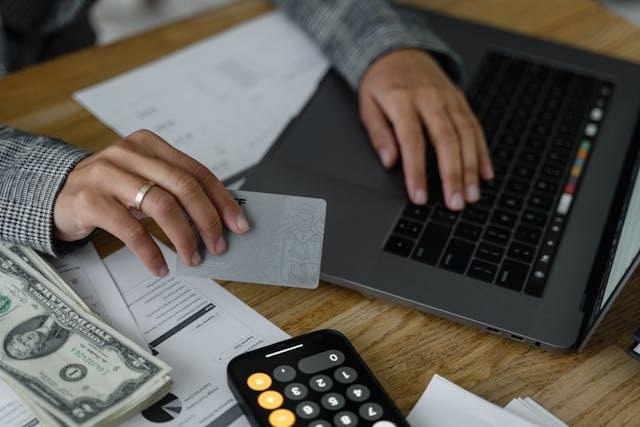 Person holding a bank card with one hand and using a laptop with the other while money, papers, and a calculator are on the desk around them